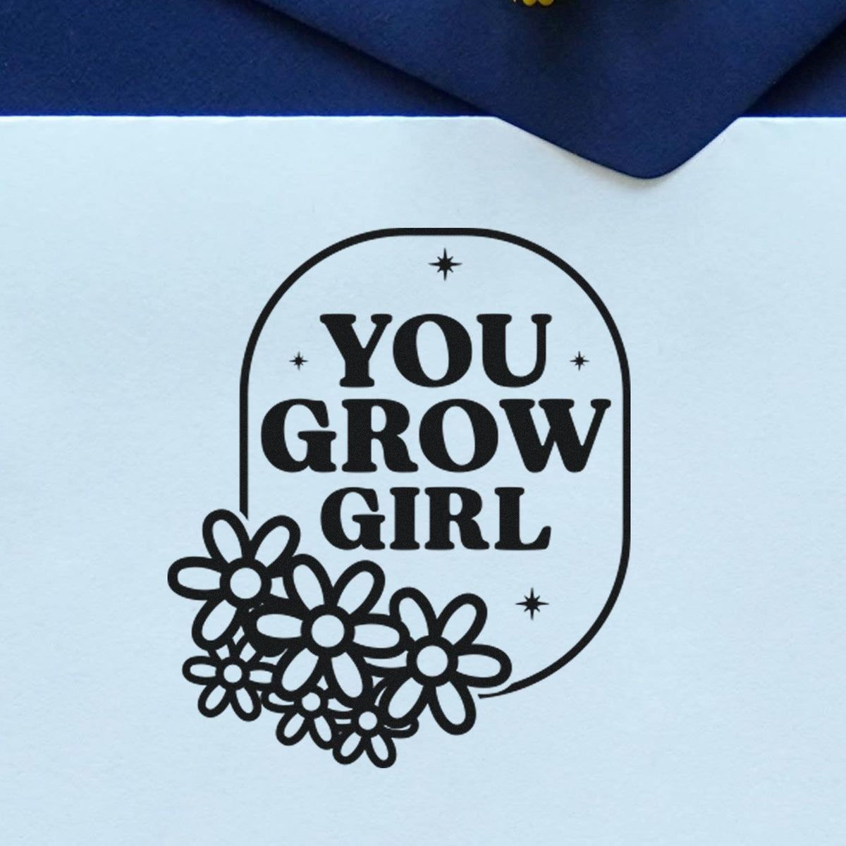 Black stamp seal with YOU GROW GIRL in bold letters and flower accents on white paper with navy background.