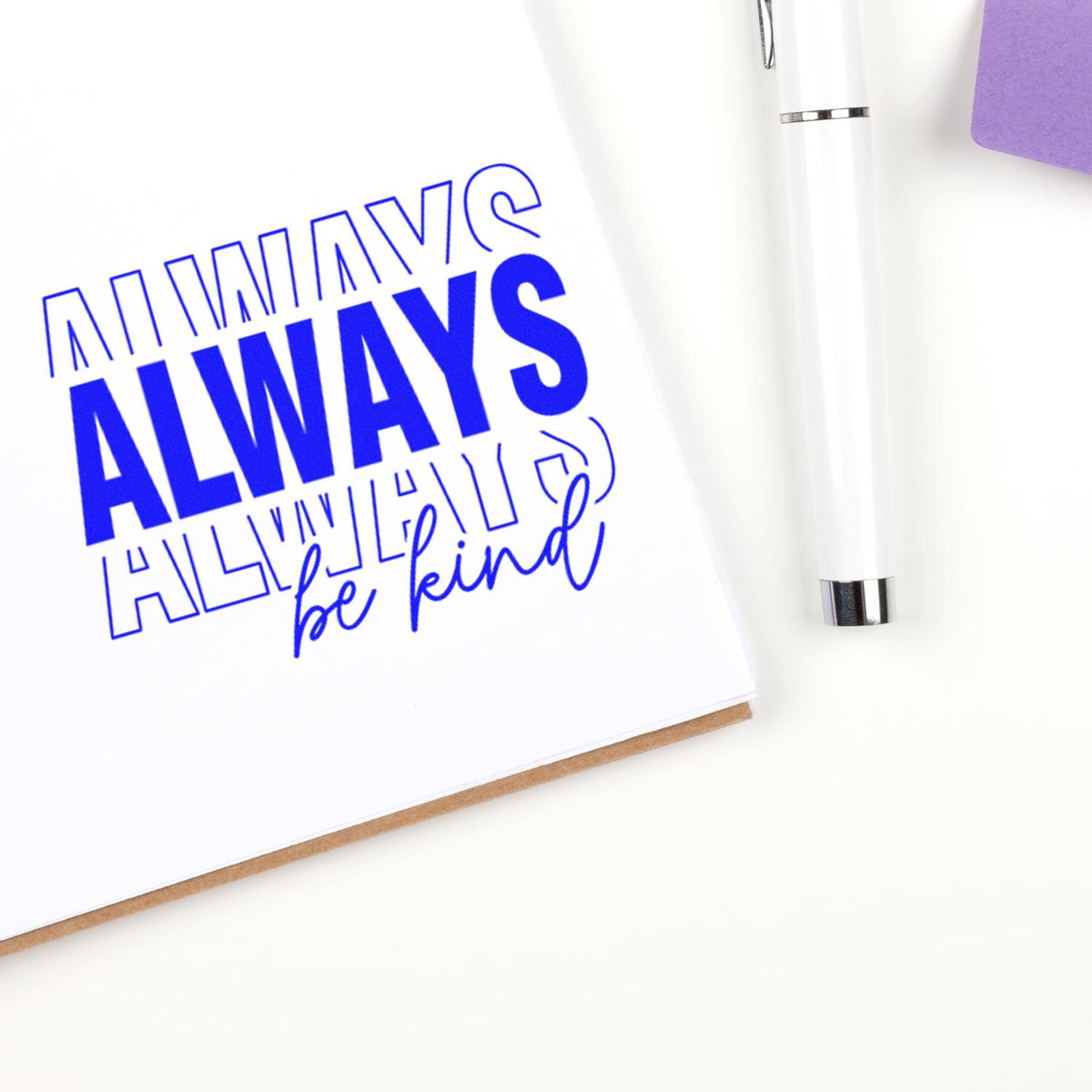 Blue stamp seal reading ALWAYS be kind on white paper, beside a white pen and purple accent.