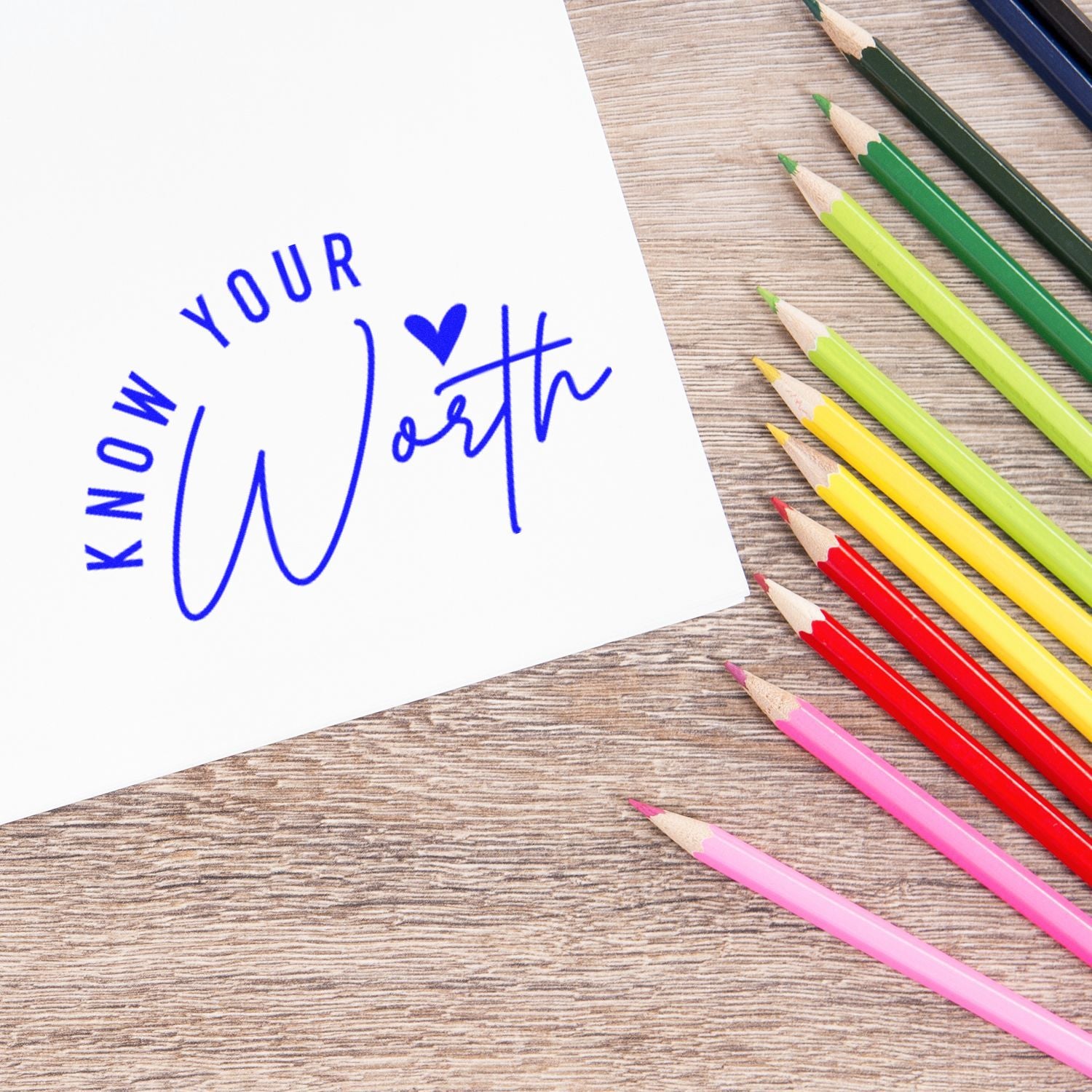 Blue Know Your Worth stamp seal on white paper beside colorful pencils on a wood desk.