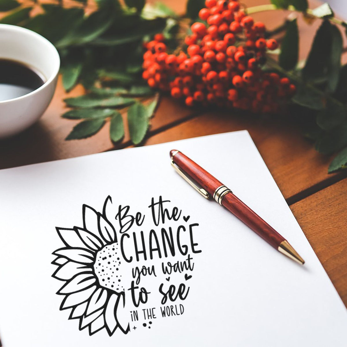 Custom stamp and seal design with sunflower quote in black on white paper, beside a burgundy pen, coffee cup, and red berries.