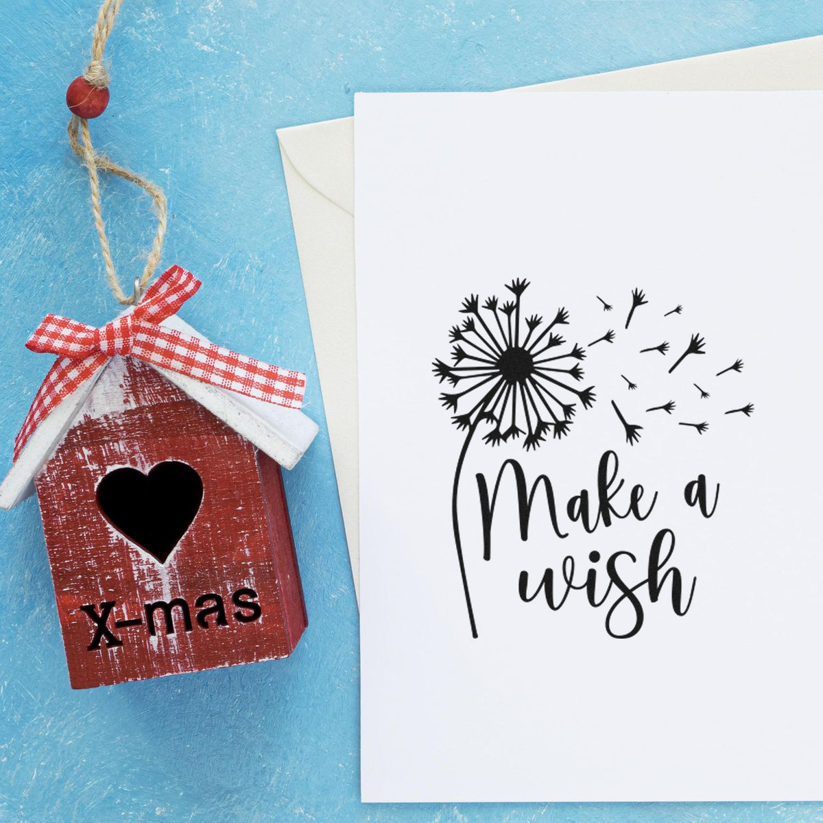 Red X-mas birdhouse stamp seal with heart cutout and plaid ribbon beside a white Make a wish dandelion card on blue background.
