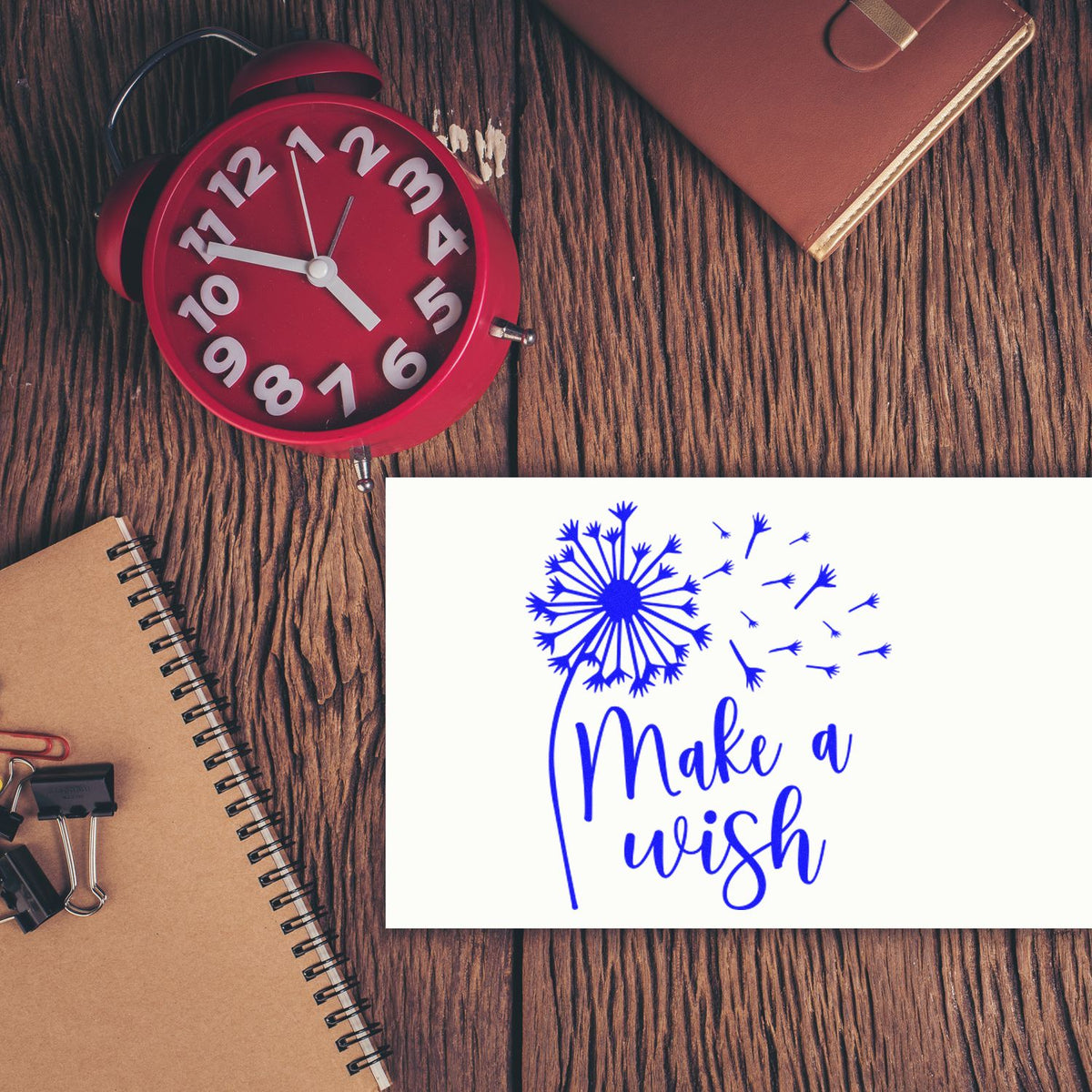 Blue dandelion Make a wish stamp and seal on a white card, with a red alarm clock, brown notebook, and leather wallet.
