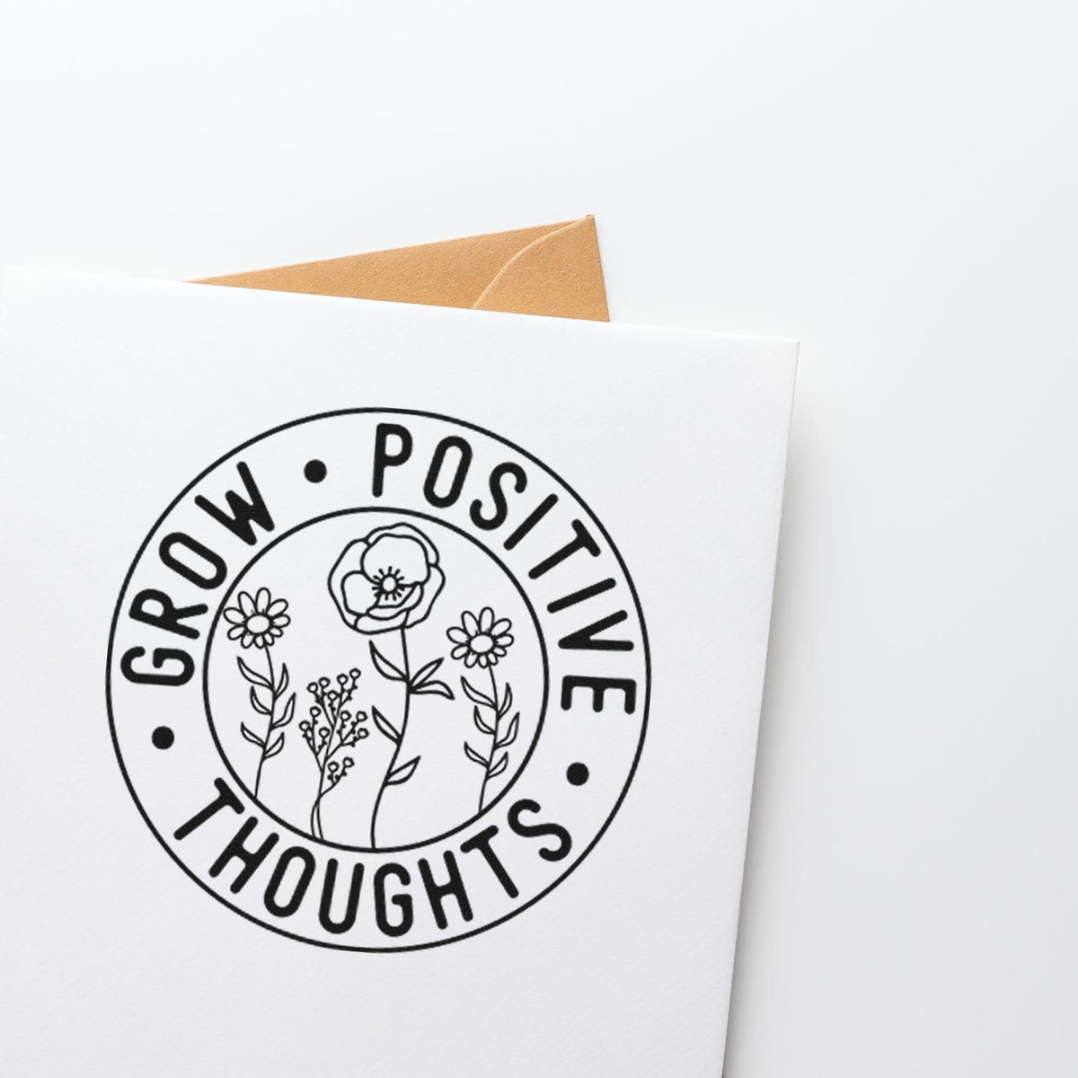Black circular stamp seal with floral line art and Grow Positive Thoughts on a white card, tan envelope behind.