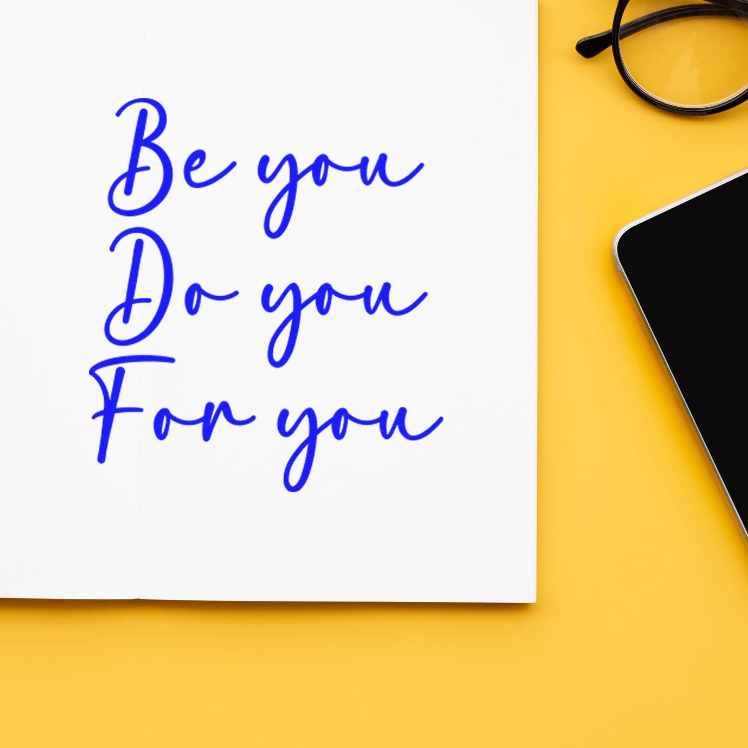 Blue custom stamp and seal reading Be you Do you For you on white paper, with black glasses and phone on yellow background.