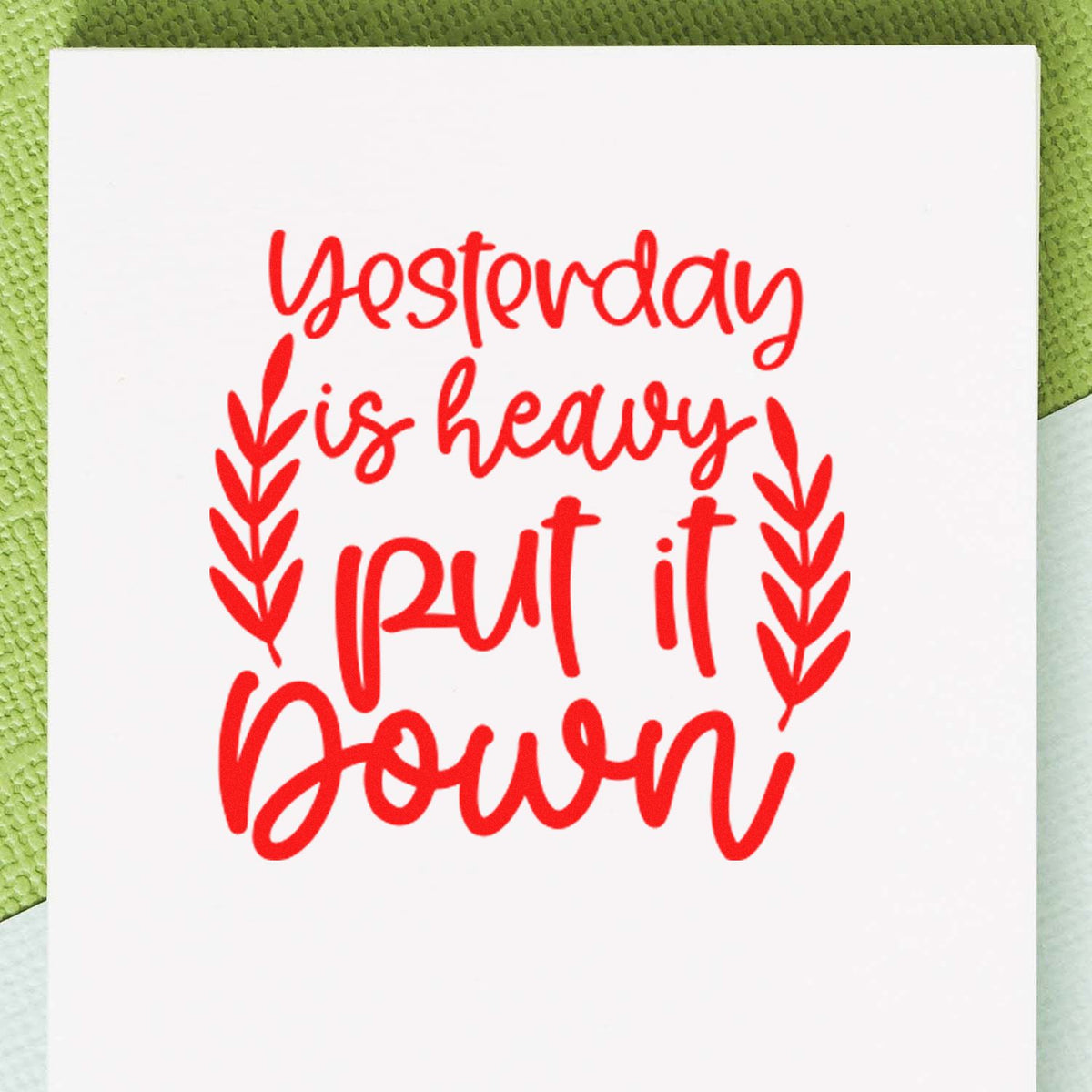 Red rubber stamp on white card reads yesterday is heavy, put it down with leafy accents on green textured background.