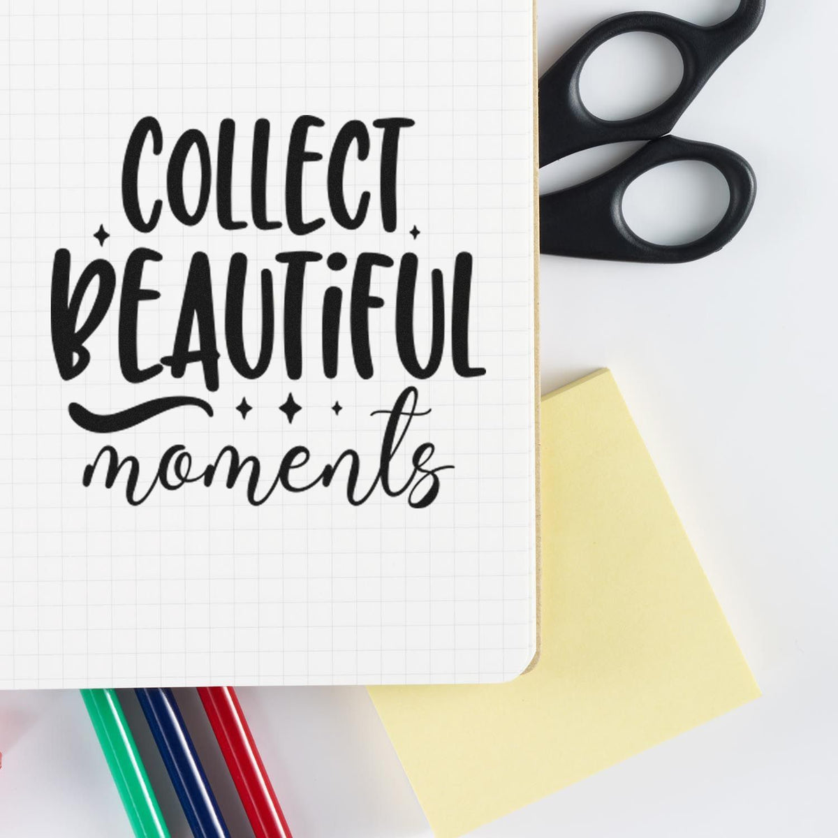 Black rubber stamp design reading Collect Beautiful moments on grid paper, with black scissors, yellow note and colored pens.
