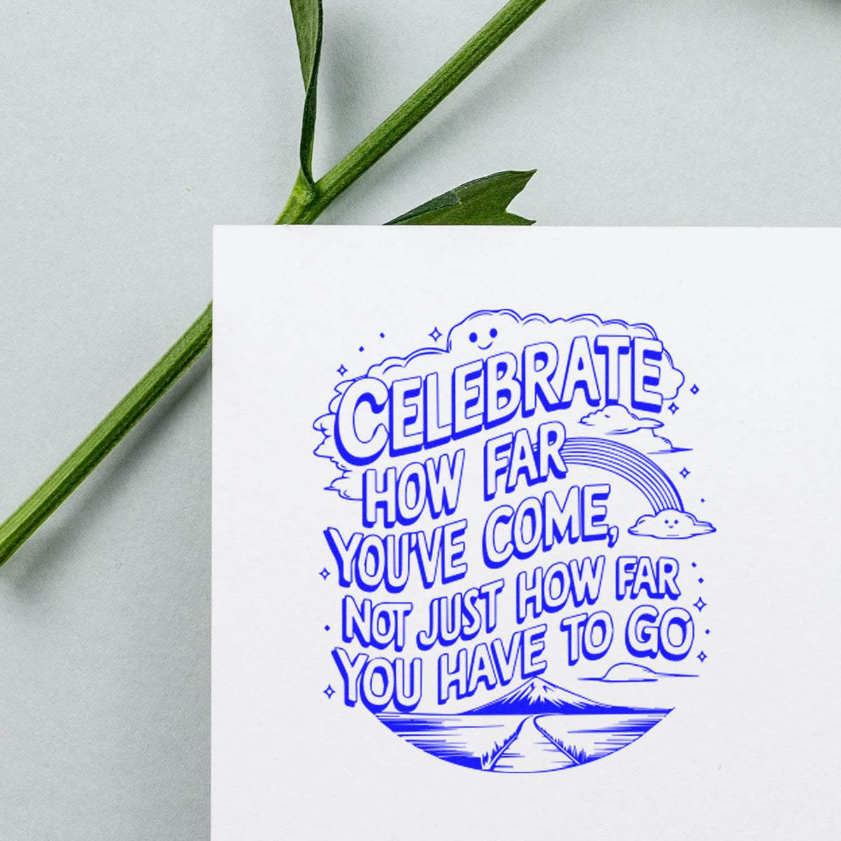 Blue celebratory stamp seal on white card with rainbow, clouds, and mountain road; green stem on pale background.