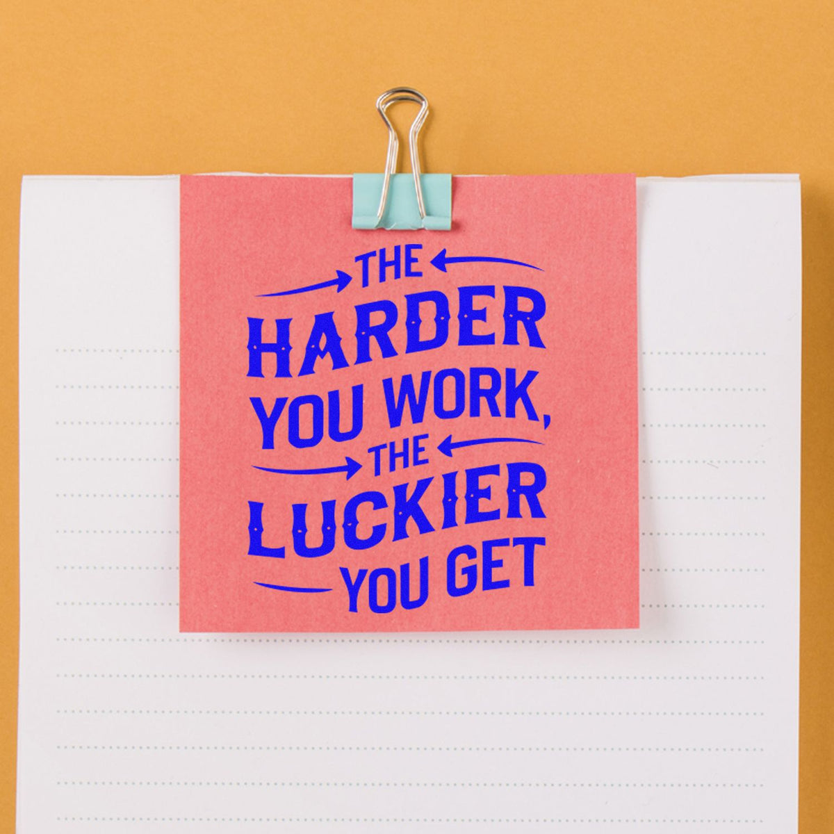 Pink memo clipped to lined notebook with blue motivational stamp reading The harder you work, the luckier you get on orange.