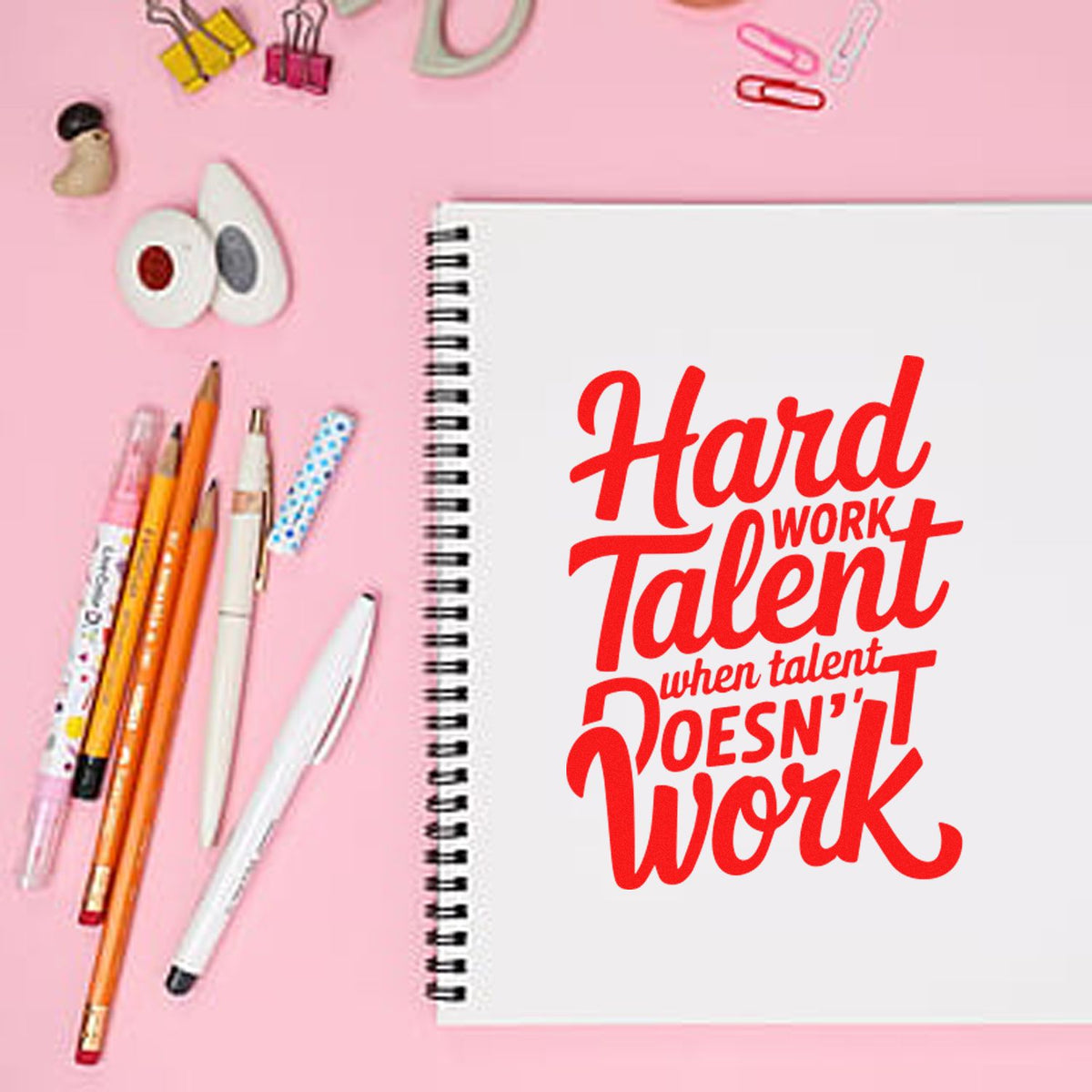 Pink flat lay with spiral notebook, red hard work quote stamp, pens, binder clips, and stamp and seal tools.