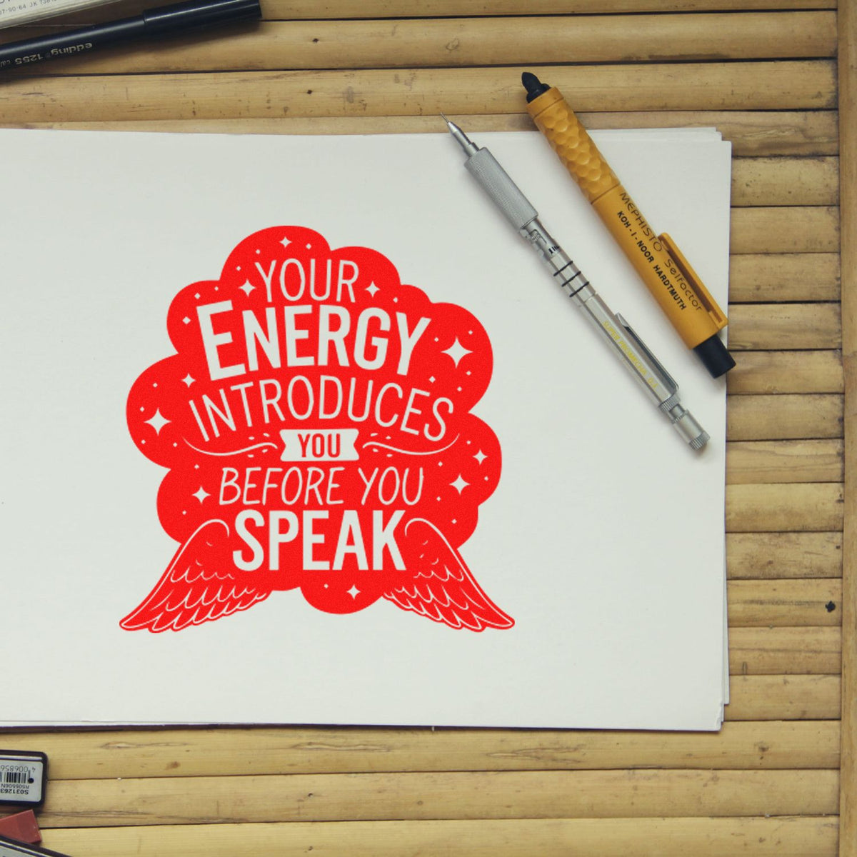 Red quote stamp/seal on white paper reading Your energy introduces you before you speak, with pens on a wood desk.