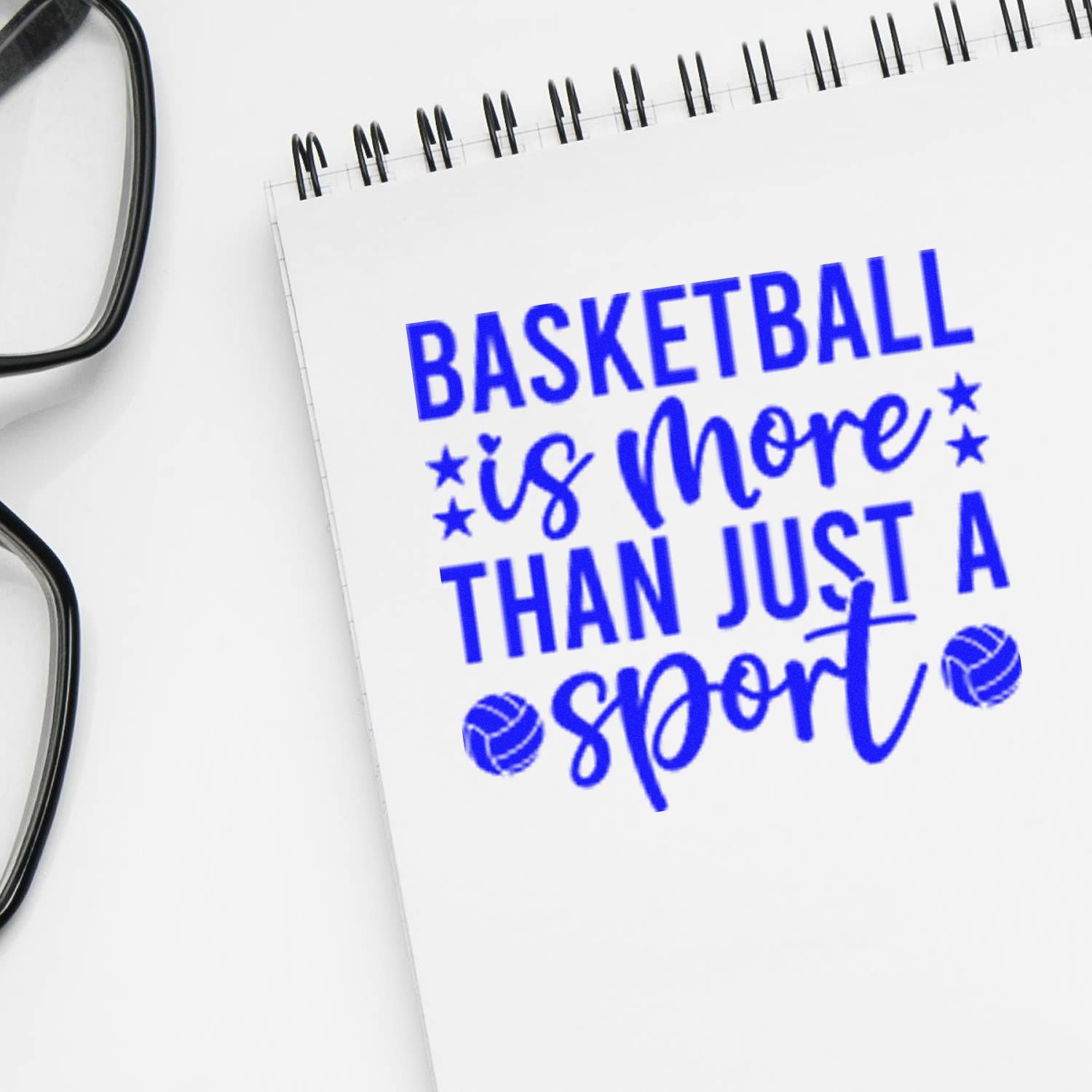 Blue basketball stamp on white spiral notebook beside eyeglasses, reading Basketball is more than just a sport.