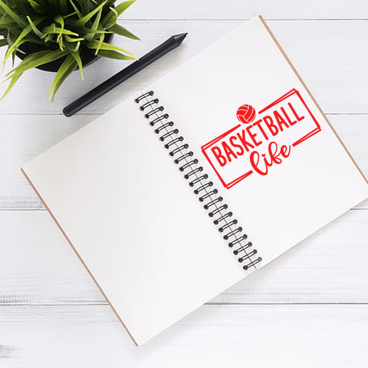 Red basketball life stamp on white spiral notebook, with black pen and green plant on a white desk.