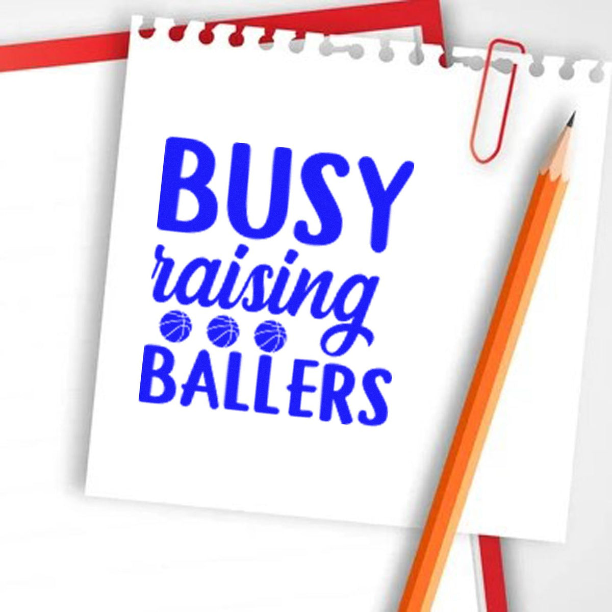 Blue custom rubber stamp reading Busy raising ballers on white notepad, with red paperclip and orange pencil.