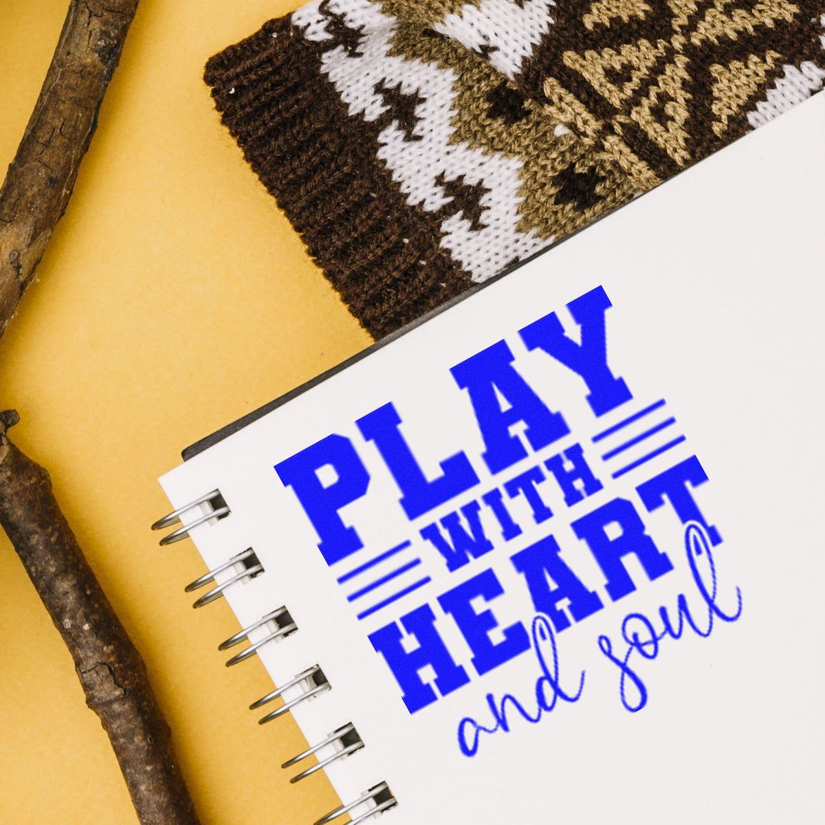 Blue stamp seal reading Play with heart and soul on a white spiral notebook, with rustic sticks and knit fabric.
