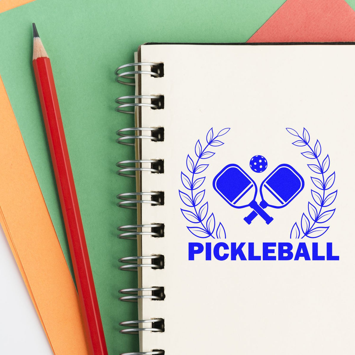 Blue pickleball stamp and seal design on a spiral notebook, with crossed paddles, leaves, and a red pencil on green and orange paper.