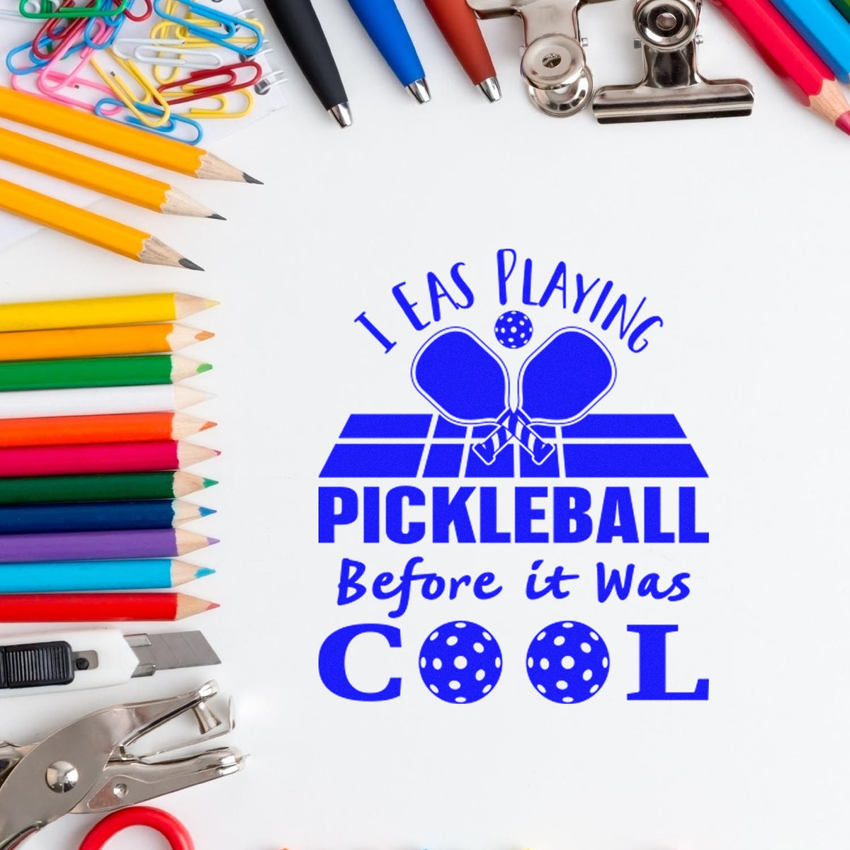 Blue pickleball stamp and seal design with colorful pencils, pens, clips, and silver paper punches on a white background.