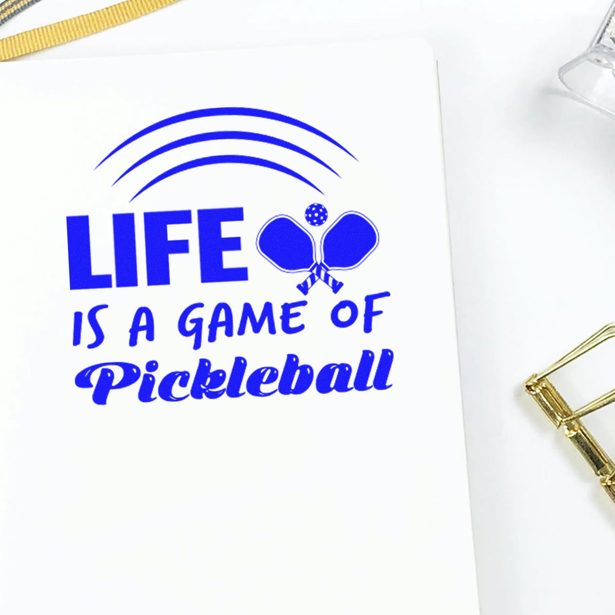 Blue pickleball stamp and seal design reading Life Is a Game of Pickleball with paddles on white paper.