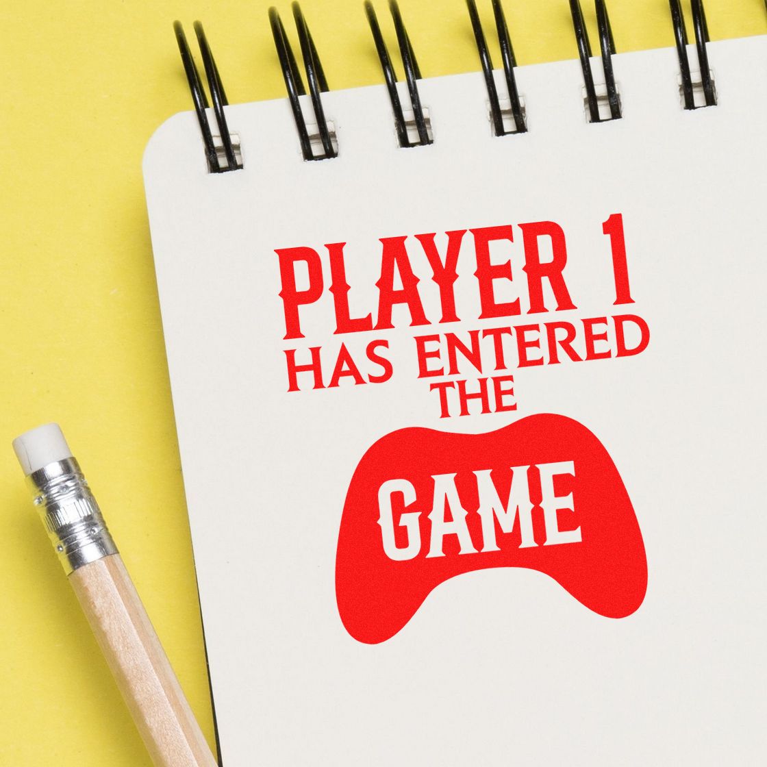 Red game-controller stamp seal reading Player 1 has entered the game on a white spiral notebook with yellow background and pencil.