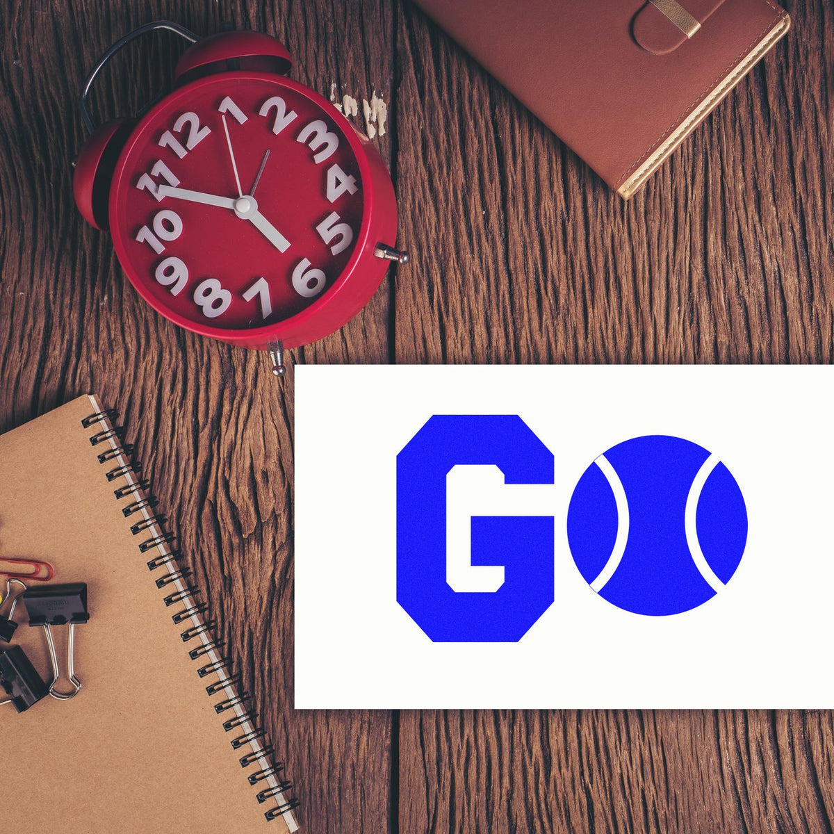 Blue GO stamp seal on white card beside red alarm clock, brown notebook, and planner on rustic wood desk.