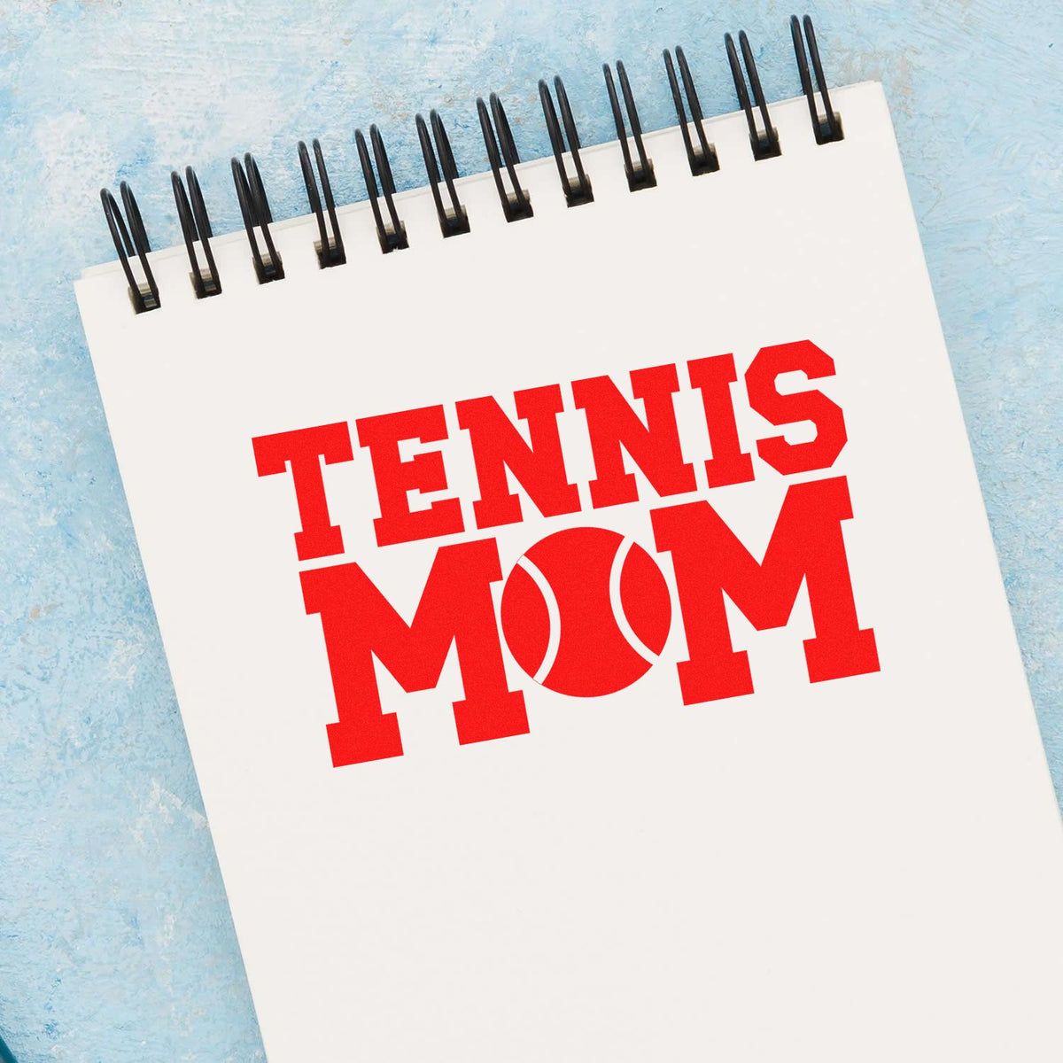 Red TENNIS MOM stamp and seal on a white spiral notebook, with a tennis ball O, against a light blue background.
