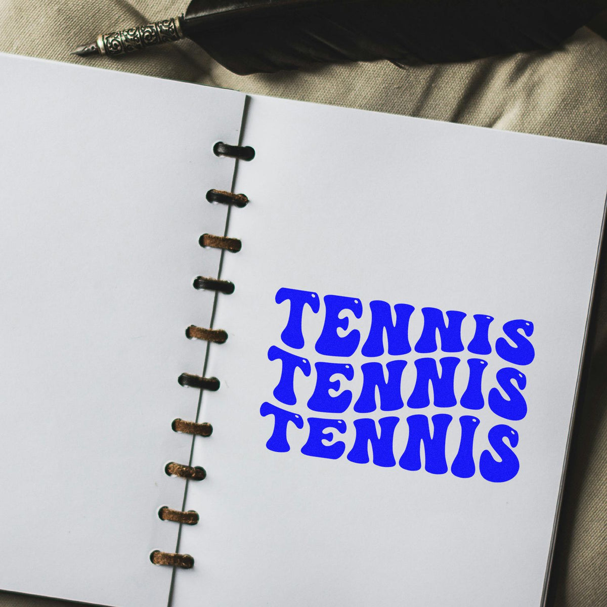Open spiral notebook with blue TENNIS stamp-style lettering, seal-like design, and a quill pen on beige fabric.