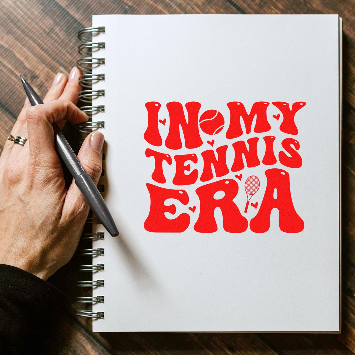 Red tennis-themed stamp/seal on a white spiral notebook, with retro lettering, hearts, tennis ball and racket.