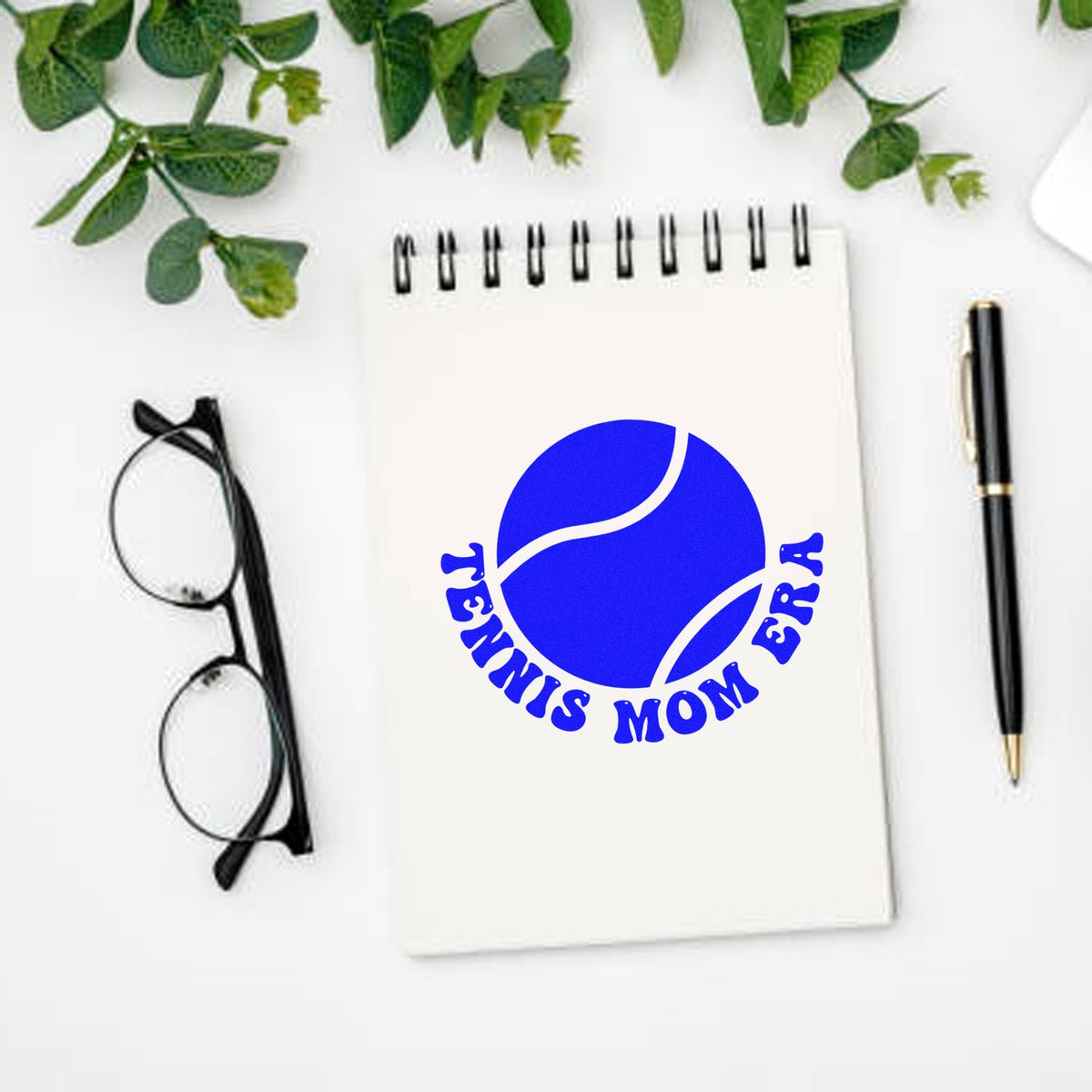 Blue tennis mom era stamp seal design on white spiral notebook, with black glasses, pen, and green leaves.