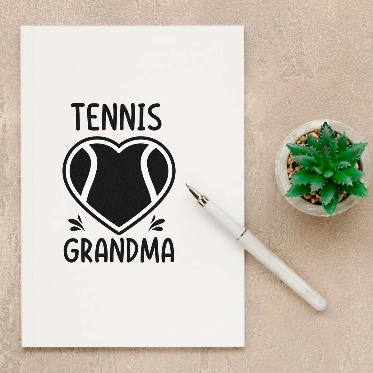 Black tennis ball heart stamp and seal design with TENNIS GRANDMA on white notebook, white pen, and green succulent.