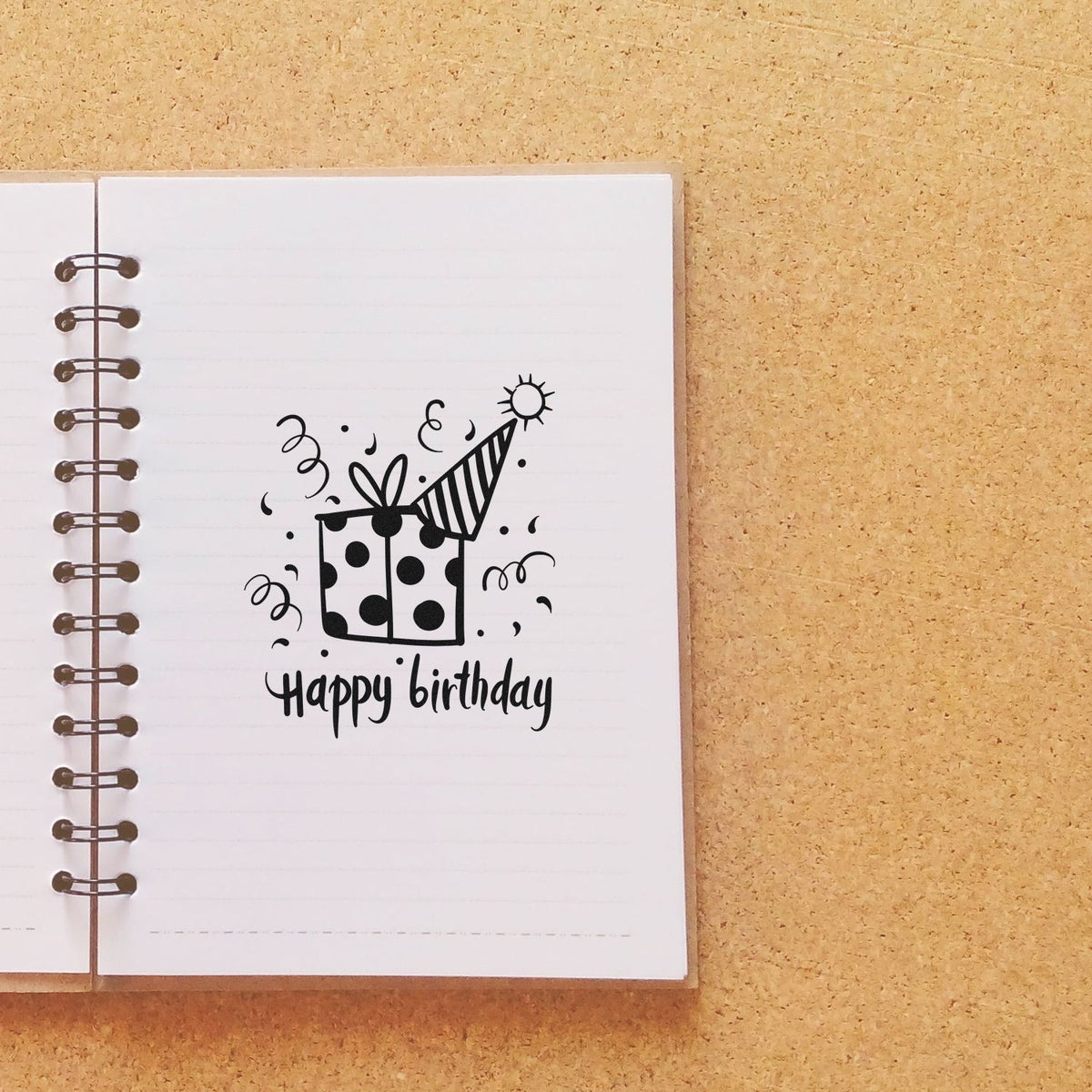 Black happy birthday rubber stamp and seal design with gift box, party hat, and confetti on a white notebook page.