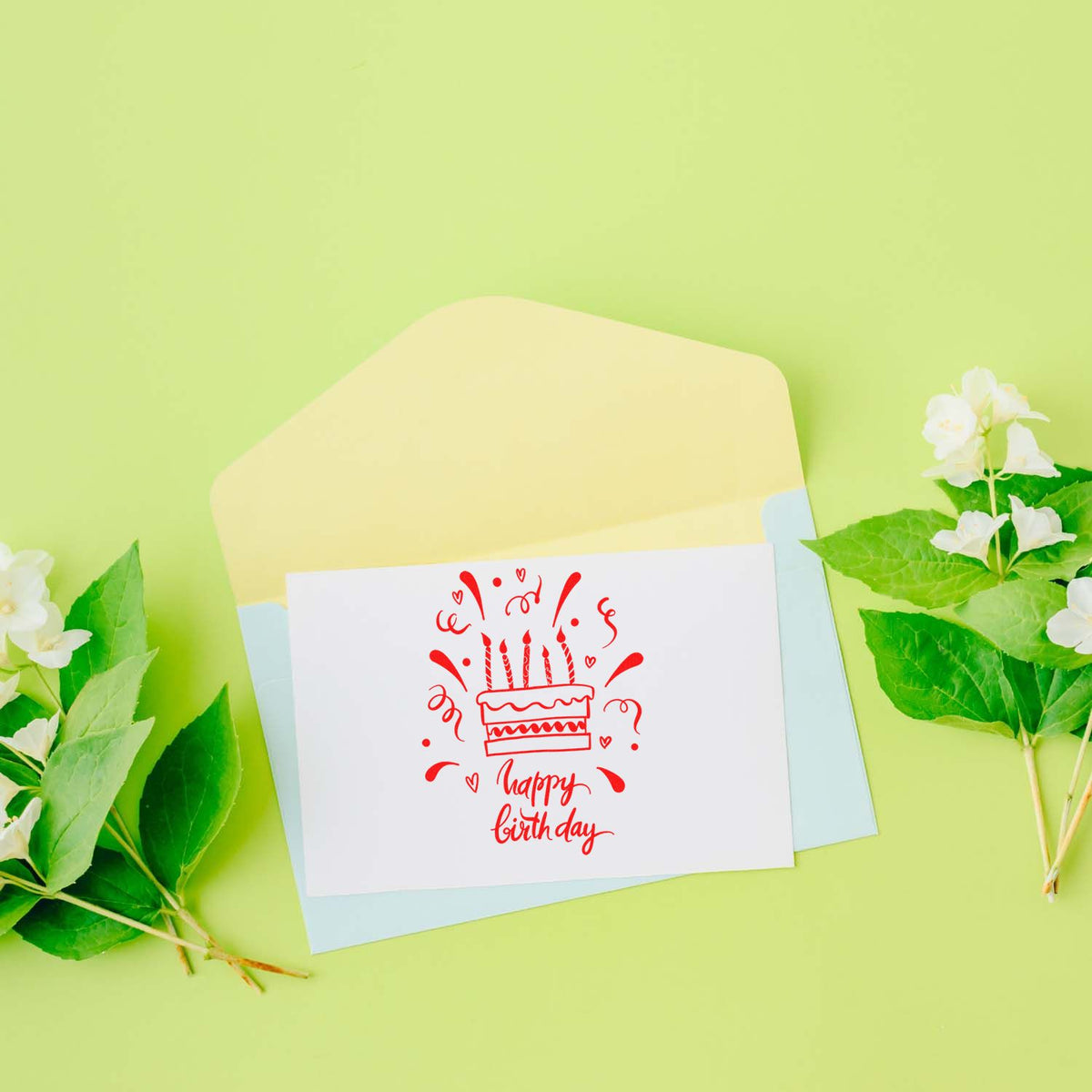 Red happy birthday cake stamp on white card with pastel envelopes, set on lime green background with white flowers and leaves