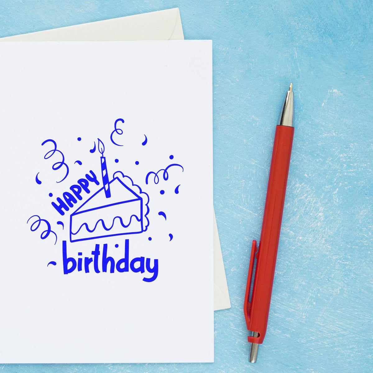 Blue birthday cake stamp on white card with confetti, beside a red pen on a light blue textured background.