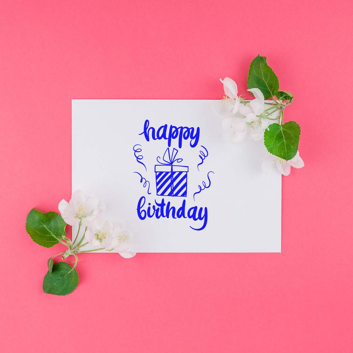 Blue happy birthday stamp with gift icon on white card, framed by white flowers and green leaves on a pink background.