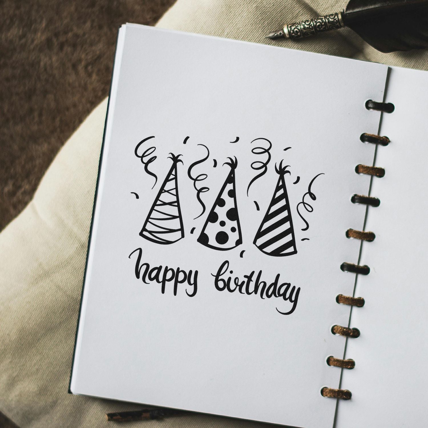 Black birthday stamp and seal design with party hats and happy birthday on white spiral-bound paper.