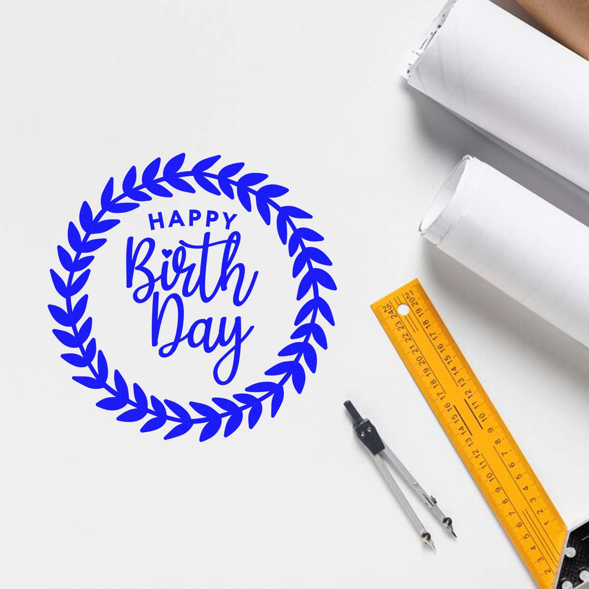 Blue Happy Birth Day wreath stamp on white paper with orange ruler, compass, and rolled paper for stamp and seal products.