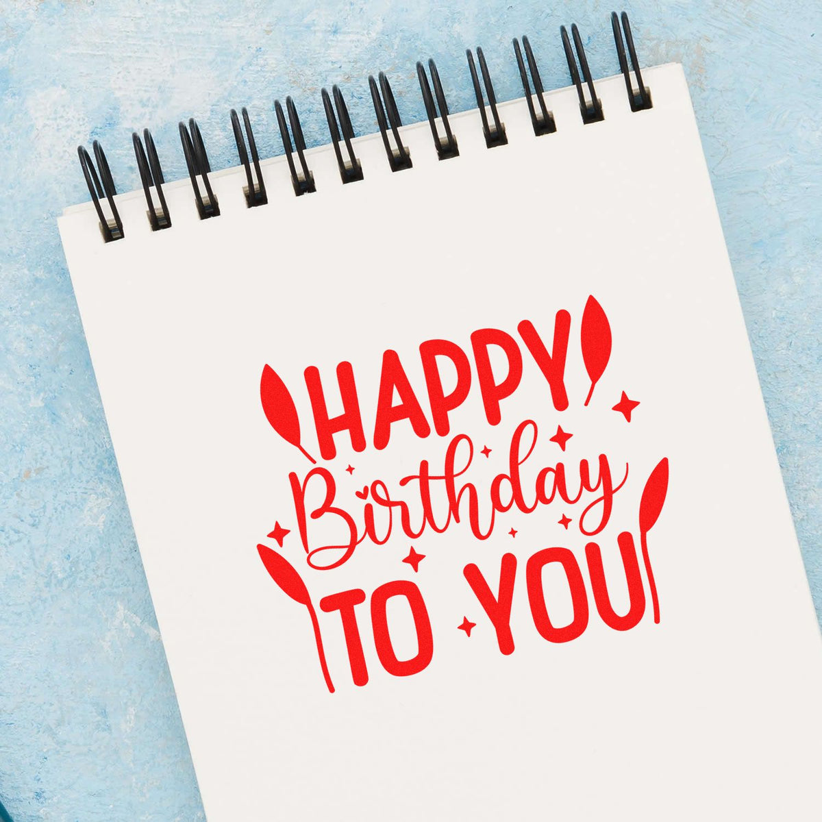 Red Happy Birthday to You stamp/seal design on white spiral notebook, with balloons and stars, on a light blue background.