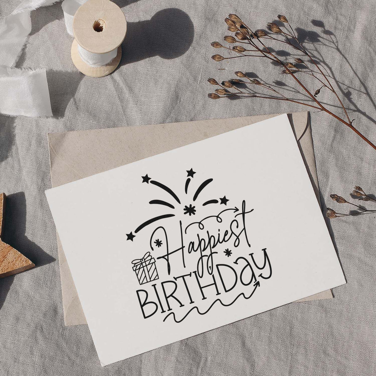 Black Happy Birthday stamp design on white card, with beige envelope, ribbon spool, and dried stems on gray fabric.