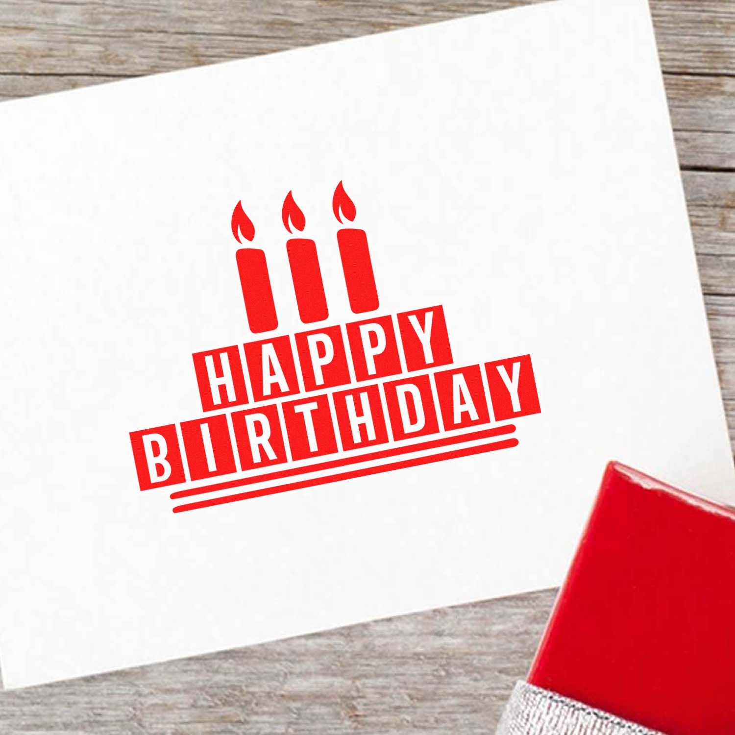Red Happy Birthday stamp with three candles on white paper, beside a red stamp pad on a wood background.