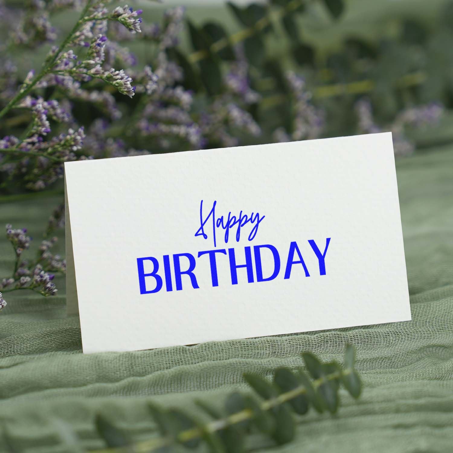 White birthday stamp and seal card with vivid blue Happy Birthday text, set among soft green foliage and purple flowers.