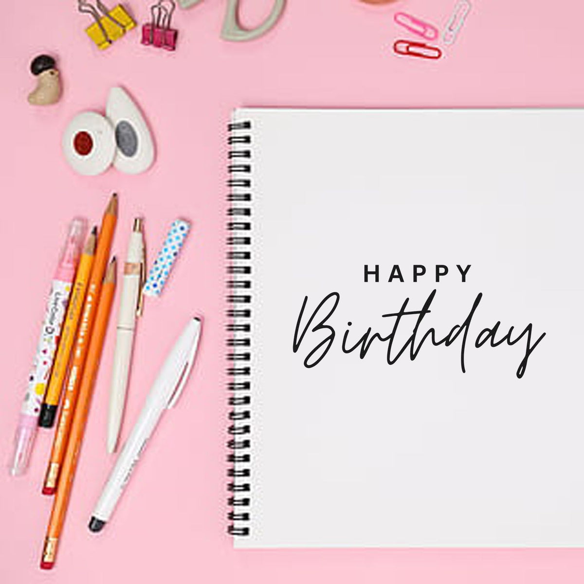 Pink flat lay of stamp and seal supplies, pens, clips, and a spiral notebook reading Happy Birthday