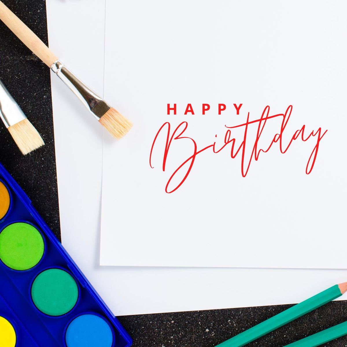 Happy Birthday stamp and seal design on white card with paintbrushes, watercolor palette, and colored pencils.