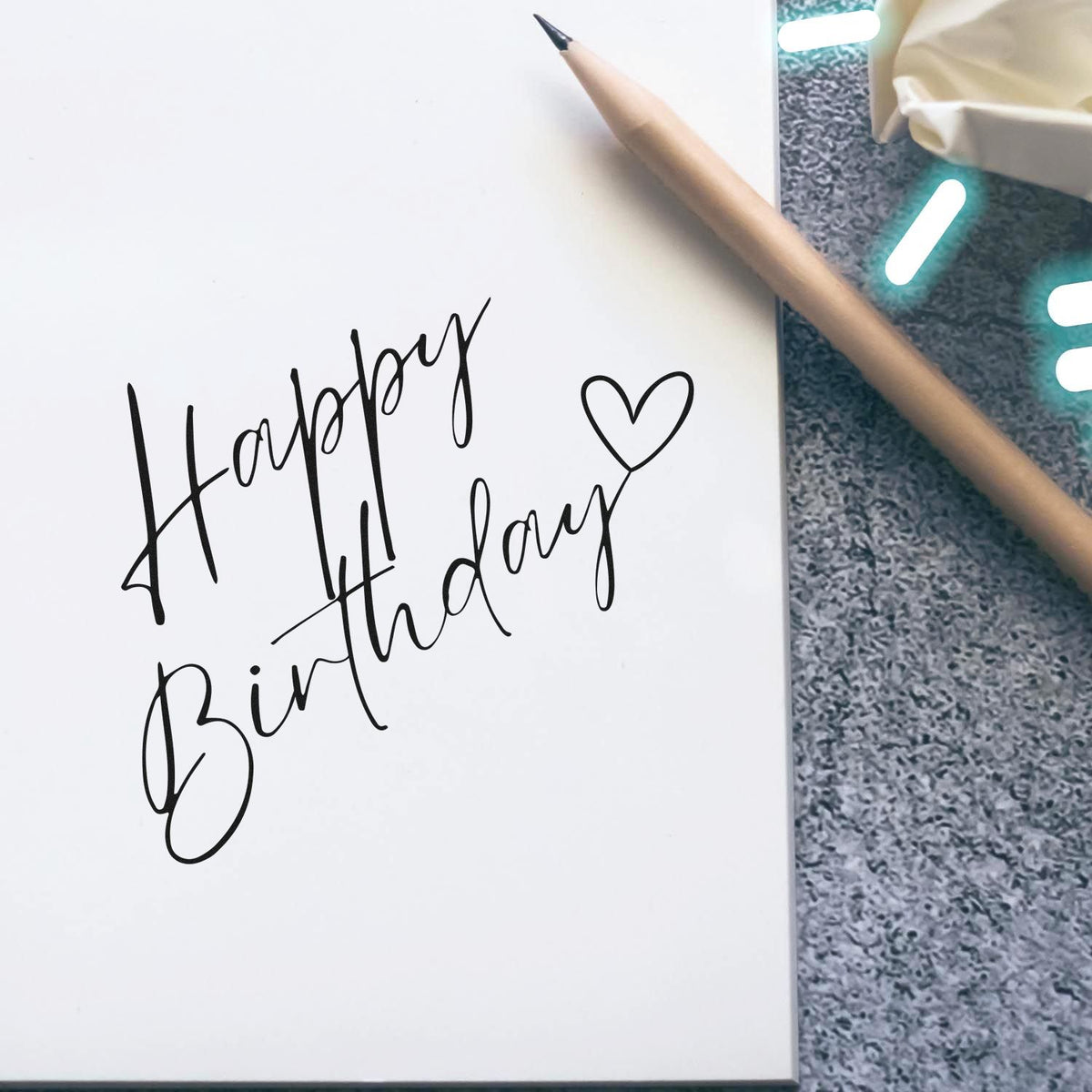 Black Happy Birthday stamp and heart seal on white paper beside a wooden pencil.