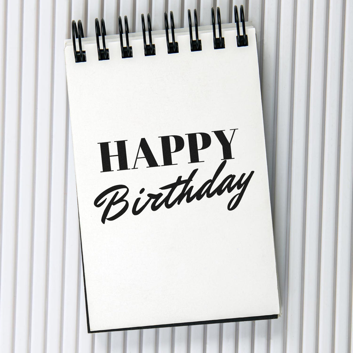 Black Happy Birthday stamp on white spiral notepad with black binding, on a light gray background.
