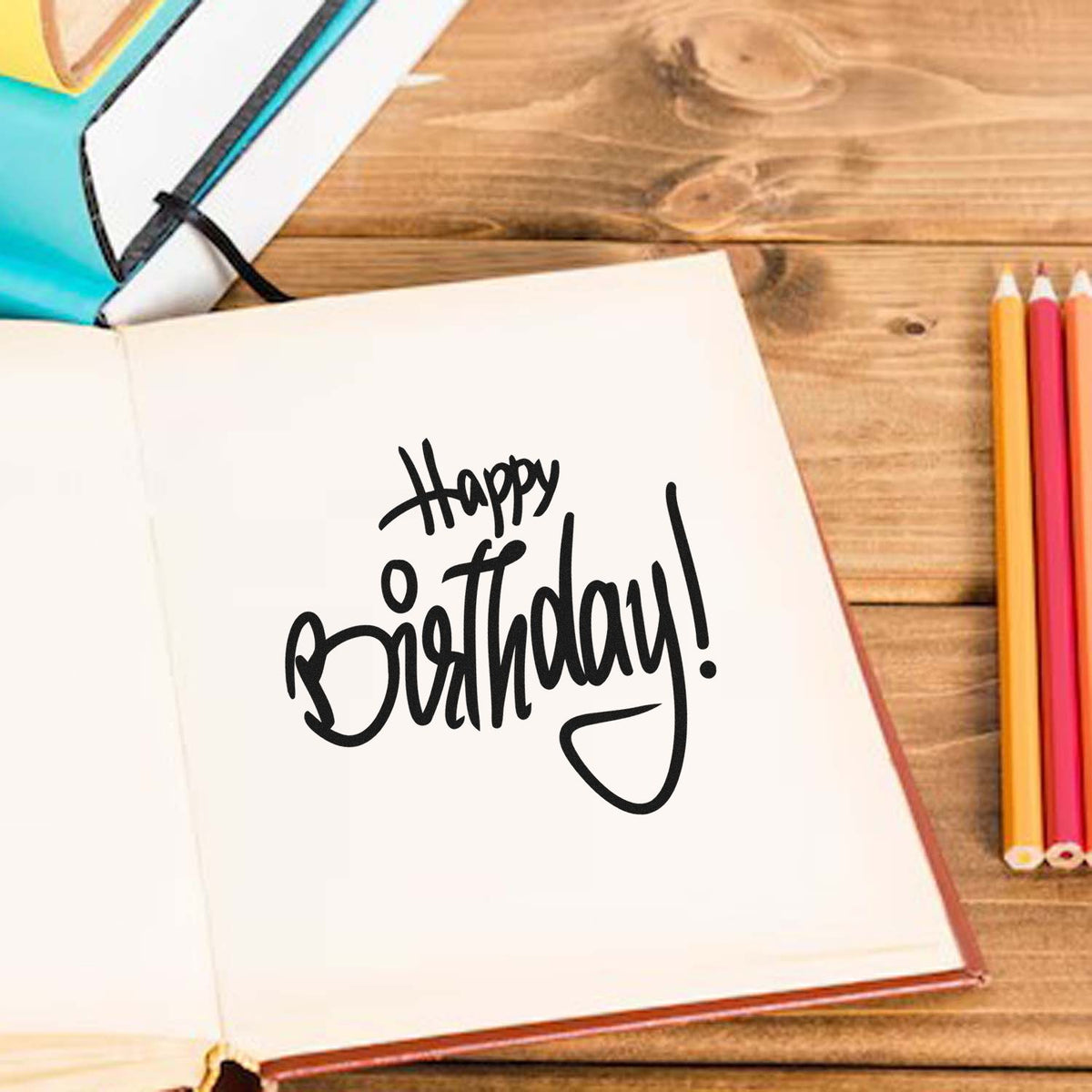 Black Happy Birthday stamp and seal on open white notebook, with colorful pencils on a wooden desk.