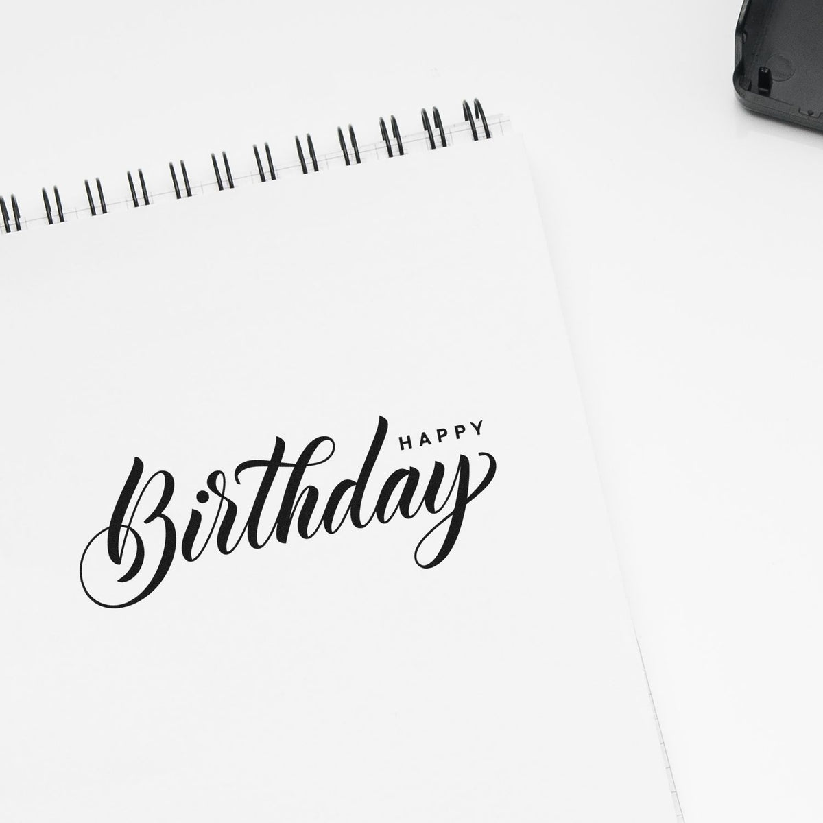 Black Happy Birthday stamp on a white spiral notebook, with a black stamp-and-seal tool in the corner.