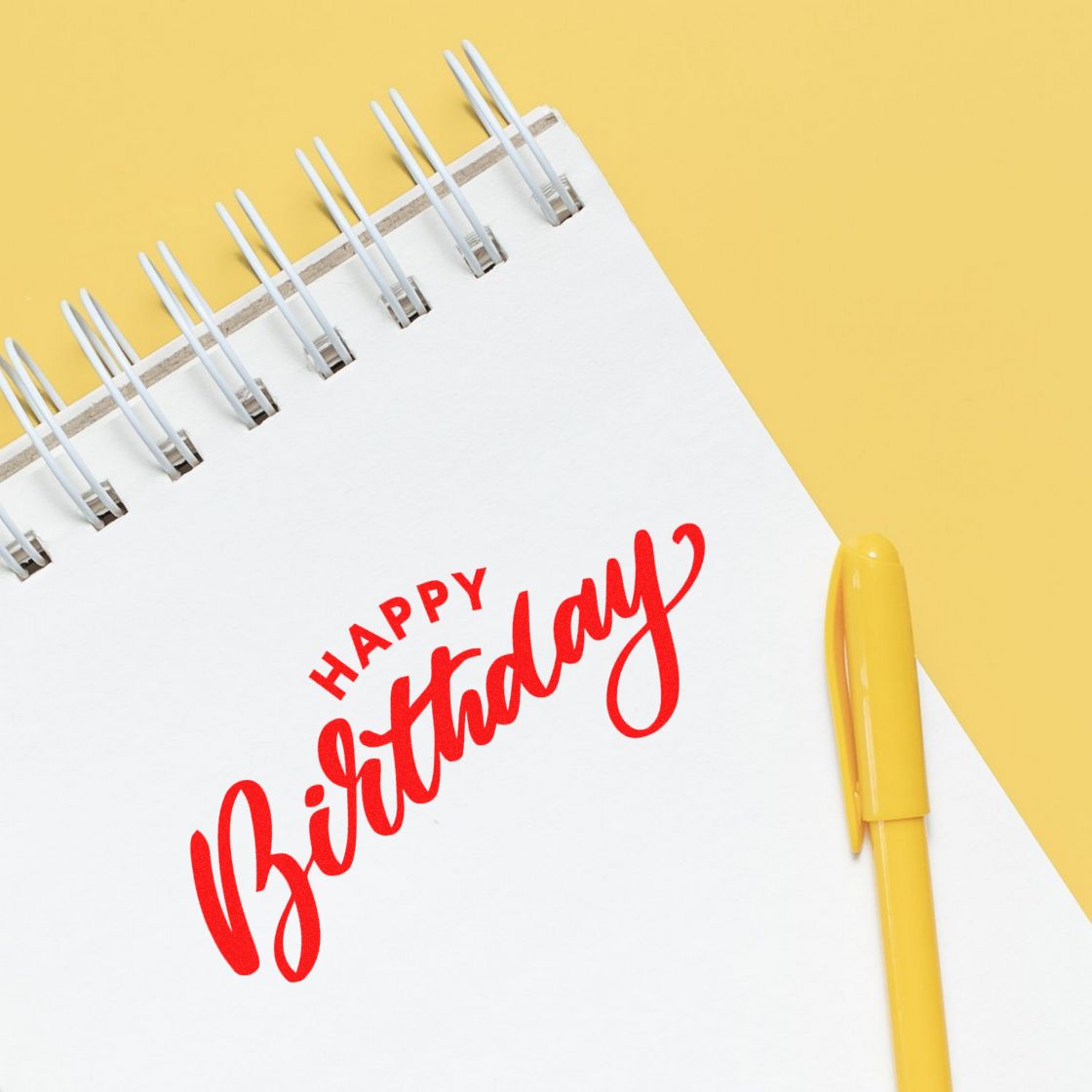 Red Happy Birthday stamp and seal on a white spiral notebook, with a yellow pen on a yellow background.