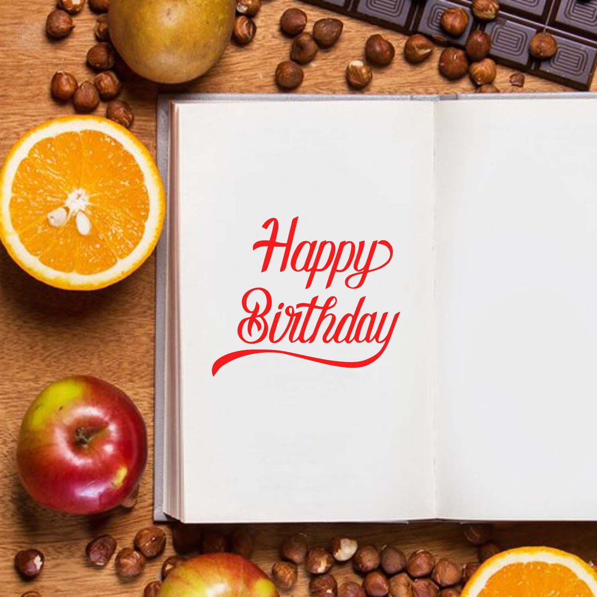 Red Happy Birthday custom stamp on open white book, surrounded by oranges, apples, and coffee beans.