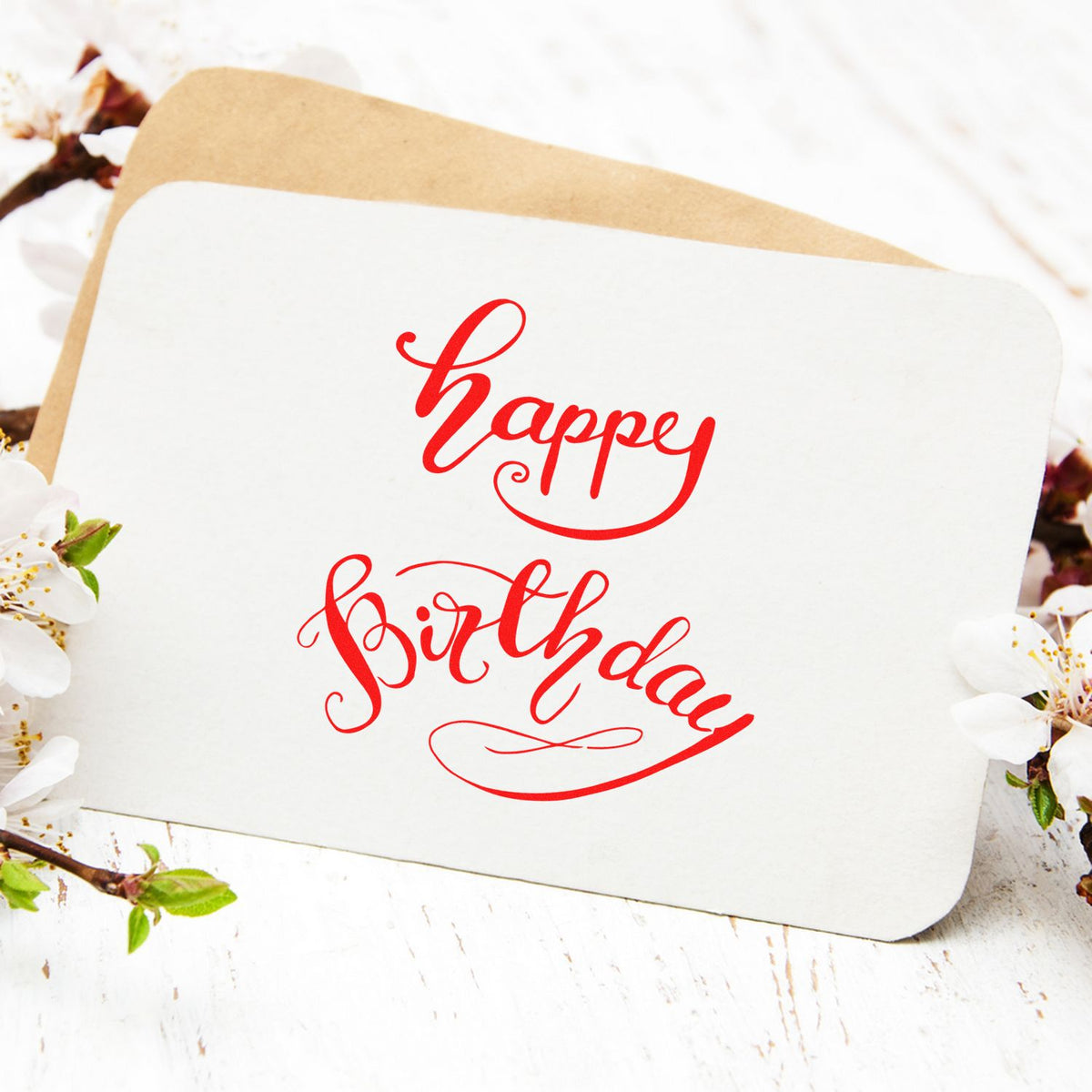 Custom stamp and seal with red happy birthday script on a white card, layered over a kraft envelope with floral accents.