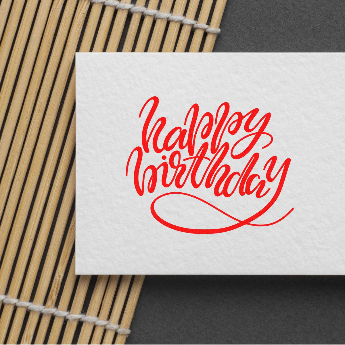 Red Happy Birthday calligraphy stamp seal on white paper, placed on a bamboo mat.