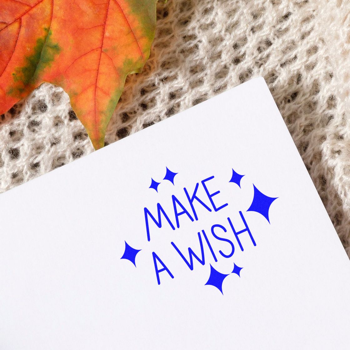 Blue Make a Wish stamp seal with starbursts on white card, beside an orange autumn leaf on beige knit.
