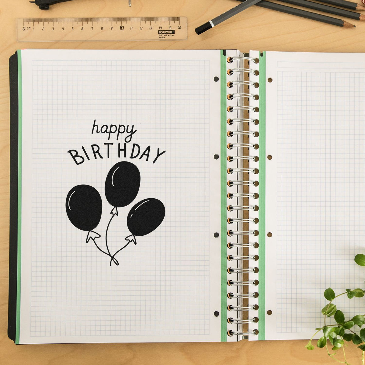 Black birthday rubber stamp with balloon design on graph paper notebook, beside a spiral-bound planner, ruler, and pencils.